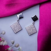 A pair of silver geometric drop earrings with a rhodium-based anti-tarnish coating, displayed on a surface with a pink scarf and white flowers in the background.