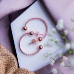 A pair of rose gold-colored semi-hoop earrings with a textured surface, displayed on a white surface with a blurred background featuring a purple satin drape and flowers.