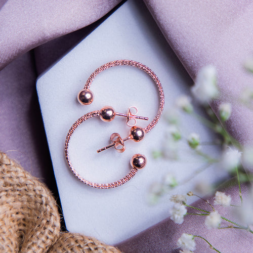 A pair of rose gold-colored semi-hoop earrings with a textured surface, displayed on a white surface with a blurred background featuring a purple satin drape and flowers.
