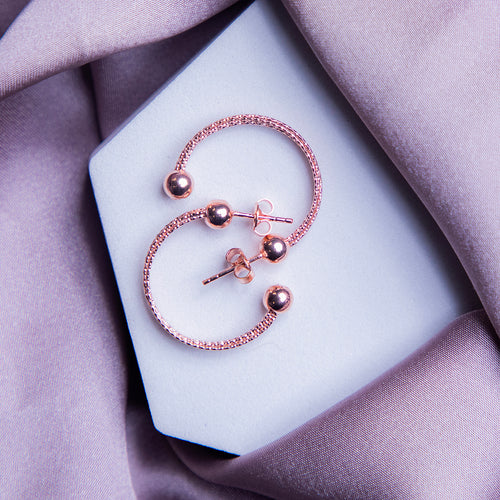 A pair of rose gold semi-hoop earrings with a textured surface, displayed on a white surface with a blurred background featuring a purple satin drape.