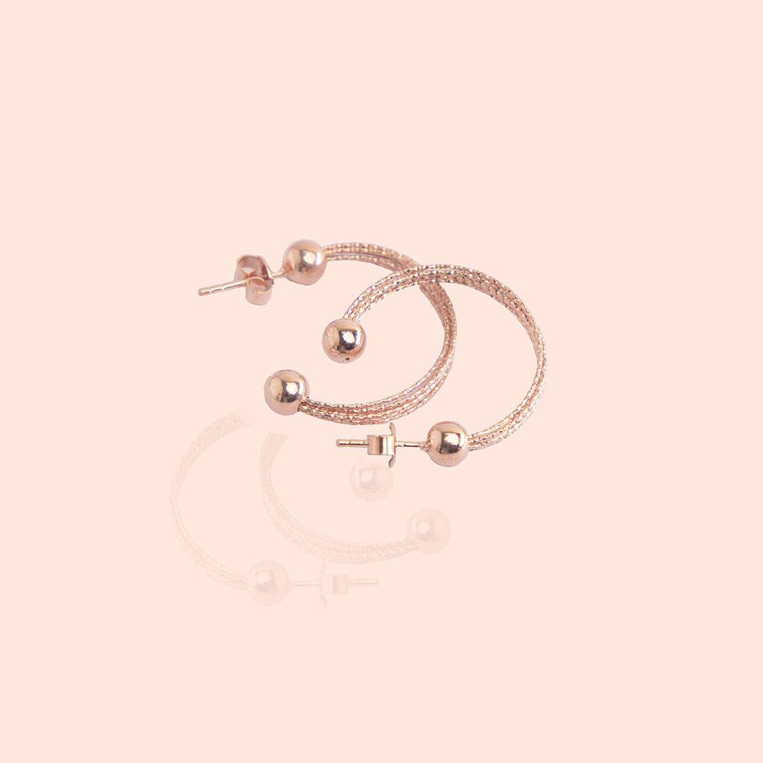 A pair of rose gold semi-hoop earrings with a textured surface, displayed on a white surface with a blurred background
