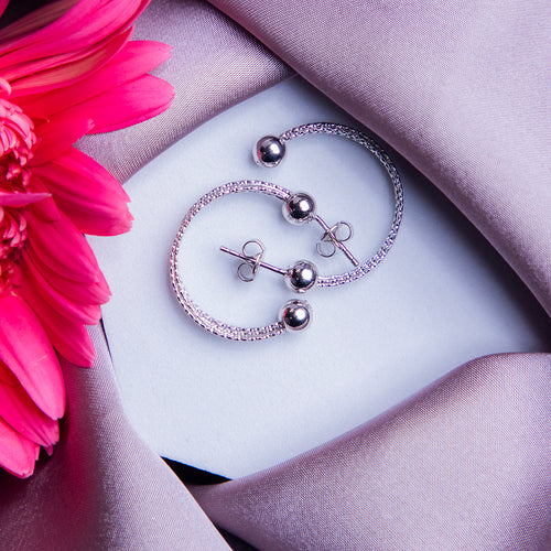 A pair of sterling silver hoop earrings with a three-layered design, displayed on a white surface next to a pink flower.
