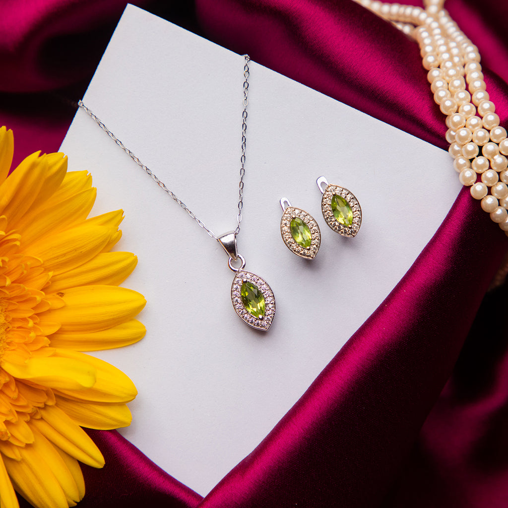 A parrot green stone pendant set with a matching pair of earrings displayed on a white card, with the pendant placed above the earrings. The set is accompanied by a yellow flower and a strand of pearls.
