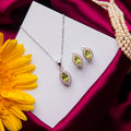 A parrot green stone pendant set with a matching pair of earrings displayed on a white card, with the pendant placed above the earrings. The set is accompanied by a yellow flower and a strand of pearls.