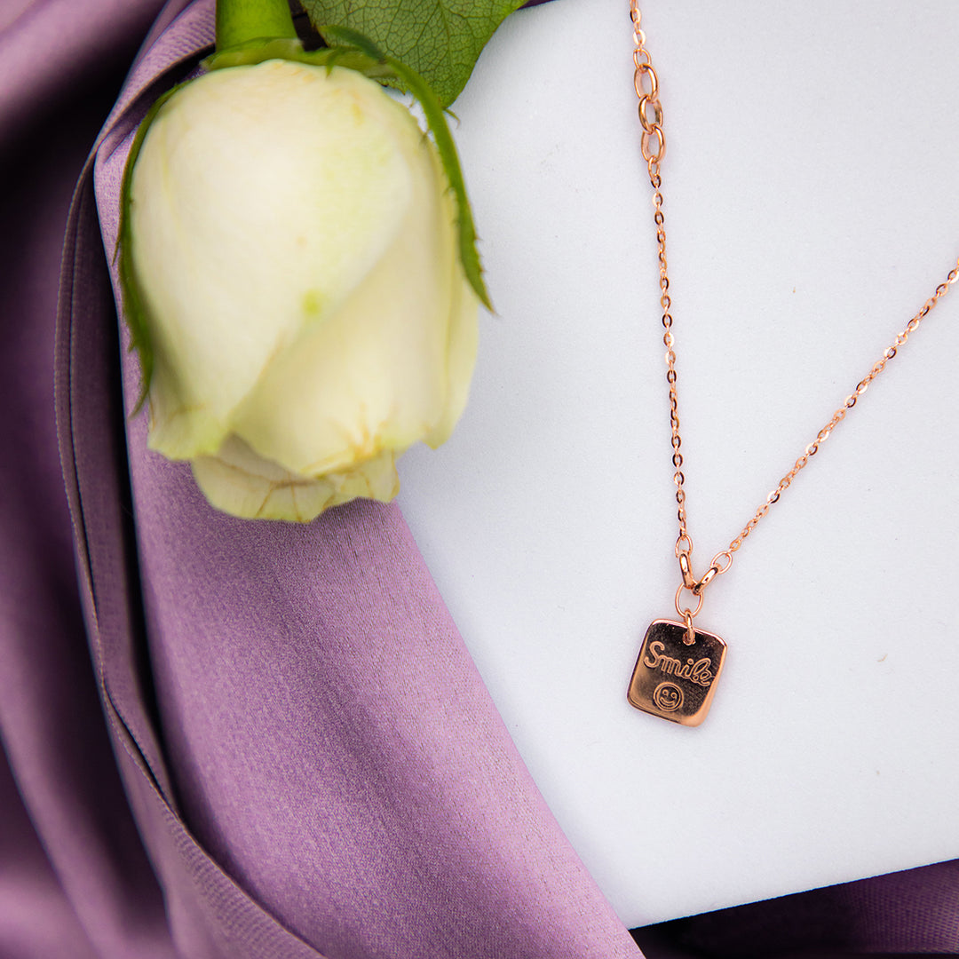 A sterling silver pendant chain with a rose gold color, featuring a smiley face design, displayed on a purple fabric background with a flower and a petal.