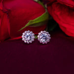 A pair of sterling silver earrings with a round white stone set in a floral pattern, displayed against a red flower background.