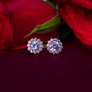 A pair of sterling silver earrings with a round white stone set in a floral pattern, displayed against a red flower background.
