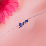 A sterling silver bracelet with the word 'Love' written in blue letters, displayed against a pink background with a pink flower.