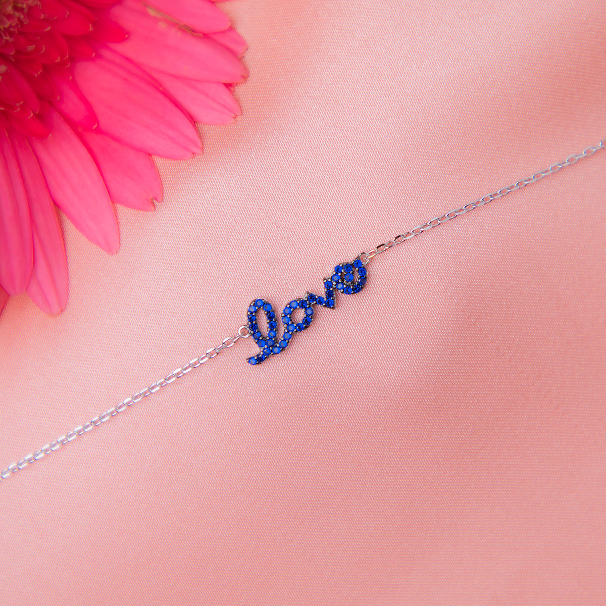 A sterling silver bracelet with the word 'Love' written in blue letters, displayed against a pink background with a pink flower.