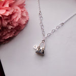 A silver pendant chain featuring a geometric bag with heart shaped charm of treasure design, displayed on a white surface with pink flower accents in the background.