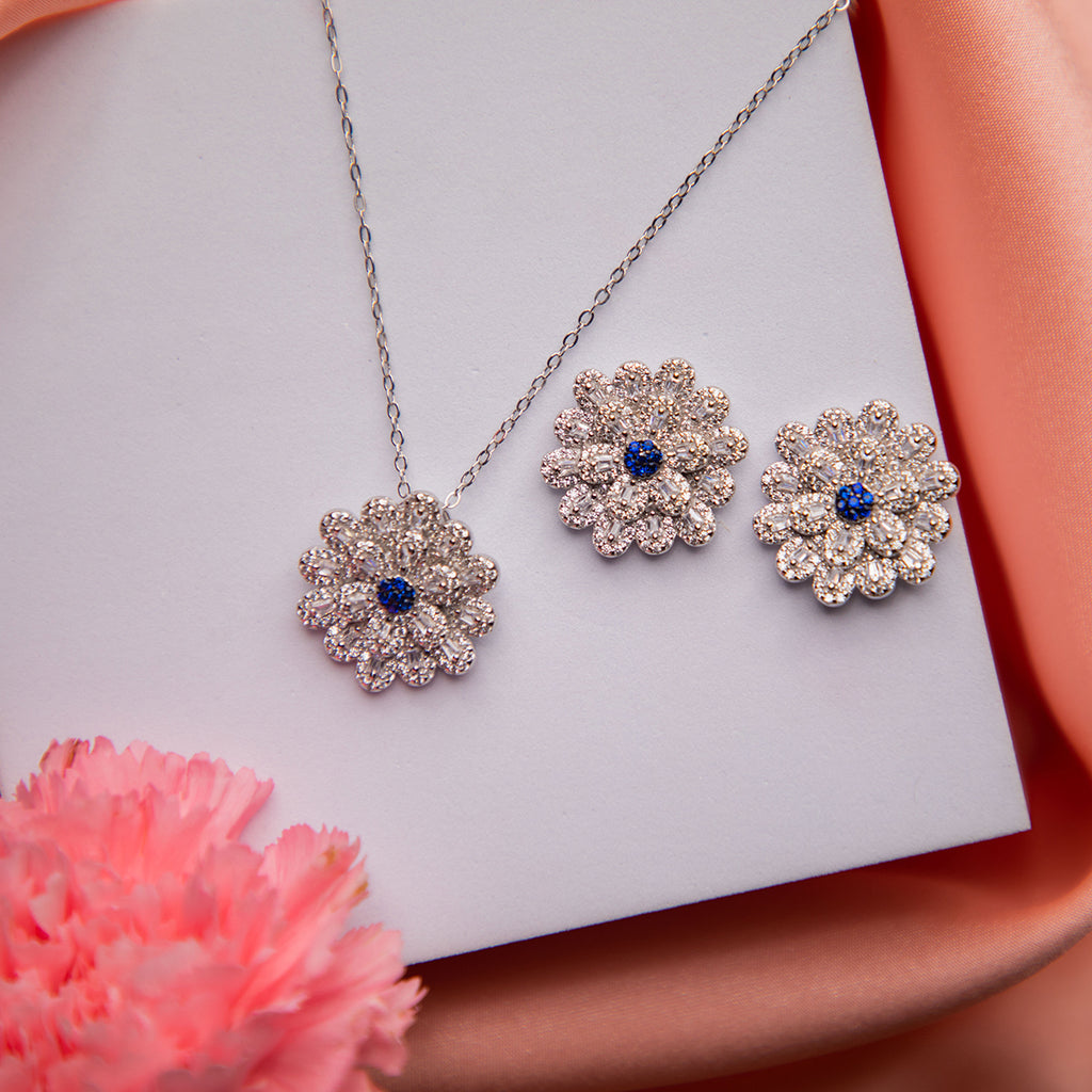 A sterling silver pendant set with a floral daisy design and blue centers, displayed on a peach fabric surface.