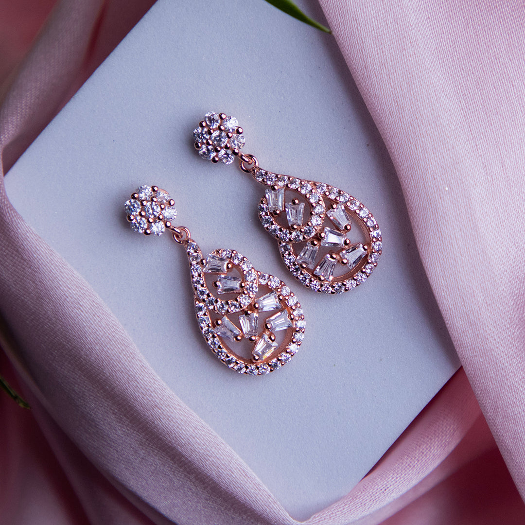 A pair of rose gold polished bejeweled statement earrings with a sparkling design, displayed on a pink surface with a white flower and a pale pink card.