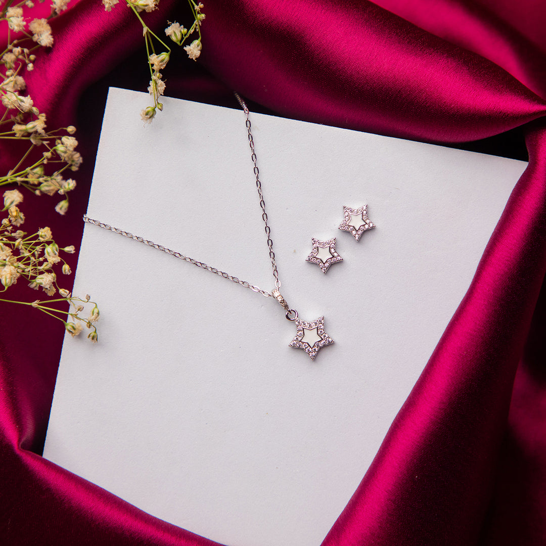 A sterling silver star pendant necklace and a pair of earrings, displayed on a white card with a pink satin backdrop and small white flowers.