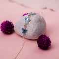 A sterling silver bracelet with blue butterfly charms placed on a rock, surrounded by purple flowers.