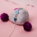 A sterling silver bracelet with blue butterfly charms placed on a rock, surrounded by purple flowers.