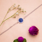 A sterling silver bracelet with a round dark blue gemstone charm accompanied by a rhodium-based anti-tarnish coating, displayed alongside floral elements.
