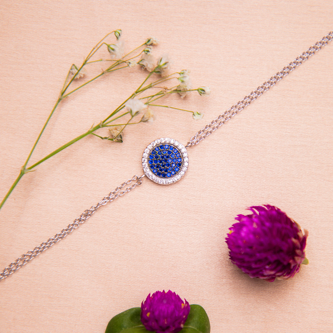 A sterling silver bracelet with a round dark blue gemstone charm accompanied by a rhodium-based anti-tarnish coating, displayed alongside floral elements.