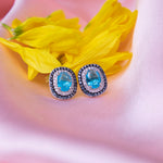 A pair of blue lagoon embellished studs earrings placed on a pink fabric along with a yellow flower petals.