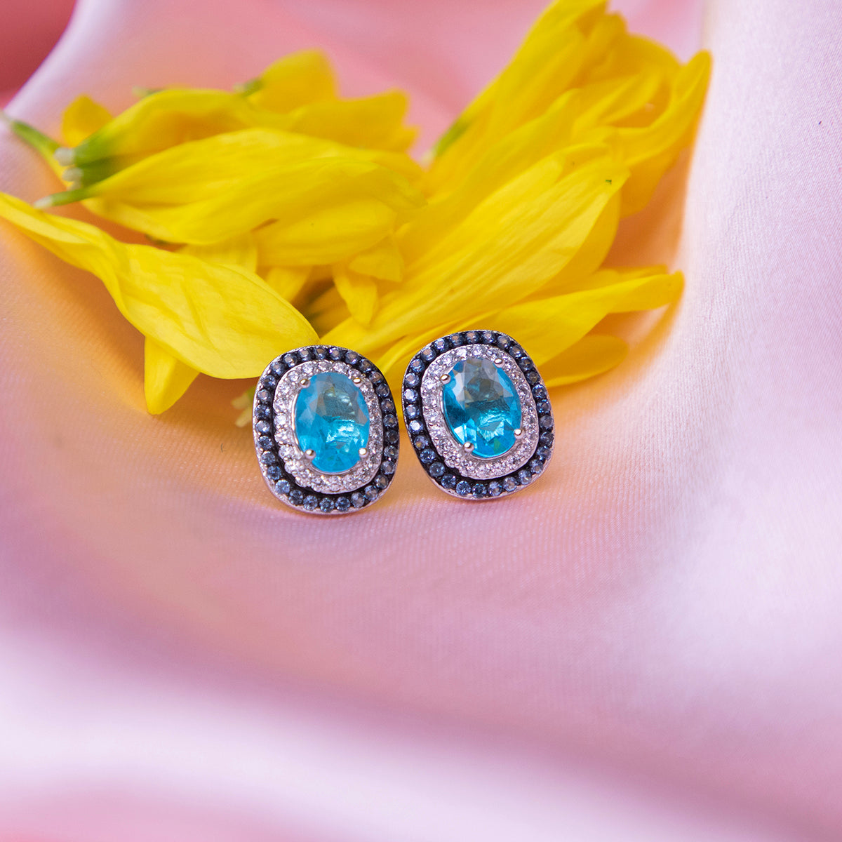 A pair of blue lagoon embellished studs earrings placed on a pink fabric along with a yellow flower petals.