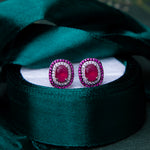 A pair of sterling silver earrings with a vibrant red gemstone centerpiece, displayed on a white surface with a green velvet background.