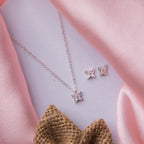 A silver pendant set with a butterfly motif and accompanying earrings, displayed on a pink fabric surface.