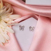 A pair of silver stud earrings in the shape of a butterfly, displayed on a pink fabric surface, with a white flower and petals nearby.