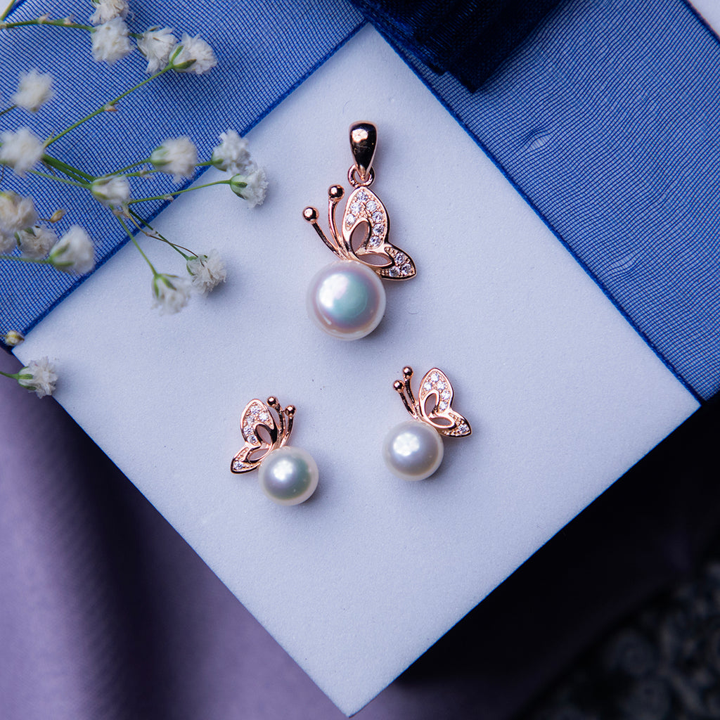 A rose gold-finish silver pendant set featuring a butterfly motif on a pearl, accompanied by matching earrings, displayed on a blue surface with white flowers.