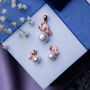 A rose gold pendant set featuring a butterfly motif on a pearl, accompanied by matching earrings, displayed on a blue surface with white flowers.