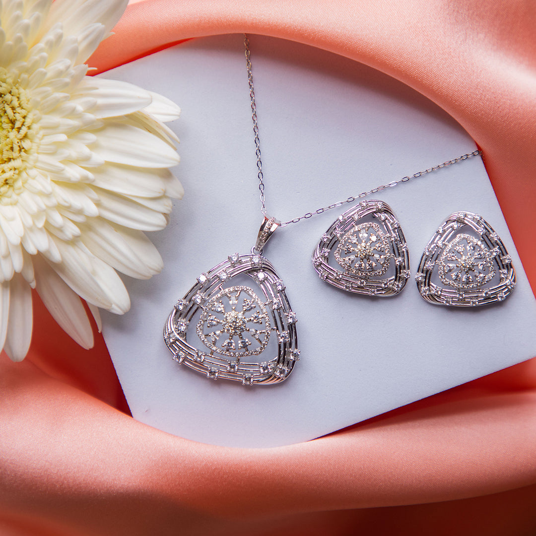 A sterling silver pendant set with a geometric pattern, consisting of a pendant and a pair of earrings, displayed on a pink fabric surface with a white flower and a card in the background.