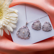 A sterling silver pendant set with a geometric pattern, consisting of a pendant and a pair of earrings, displayed on a pink fabric surface with a white flower and a card in the background.