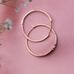 A pair of rose gold hoop earrings with a chic bangle-style design, displayed on a pink background with a floral accent.