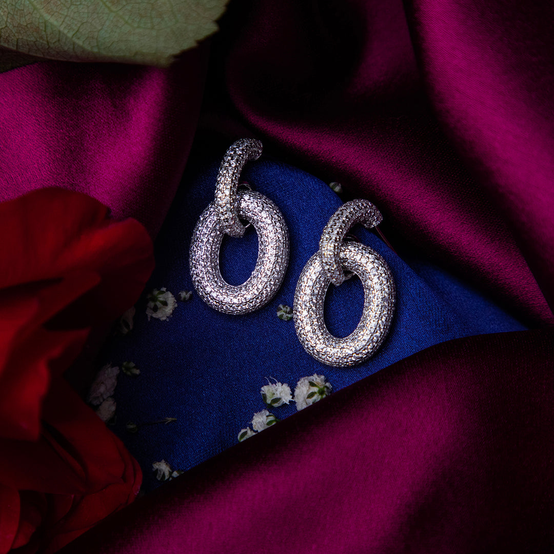 A pair of chunky silver bedazzled link earrings displayed against a purple fabric backdrop with a floral pattern and a red rose beside it.