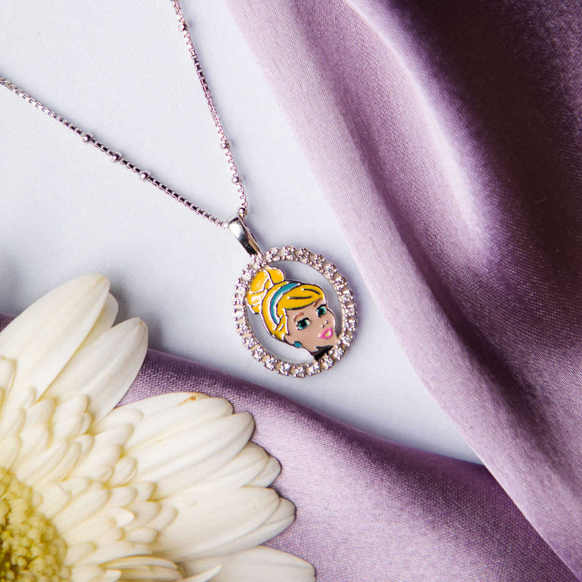 A silver pendant chain featuring a portait of Cinderella with a radiant smile, placed on a white flower against a purple backdrop.