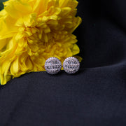 A pair of round embellished silver stud earrings placed on a dark fabric beside a yellow flower.