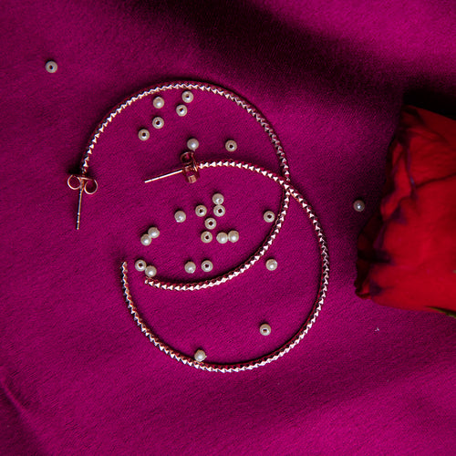 A pair of rose gold-colored sterling silver hoop earrings displayed on a purple fabric surface with scattered small silver beads and a red rose petal.