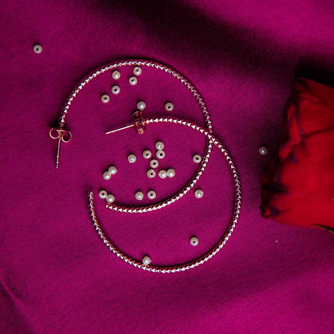 A pair of rose gold-colored sterling silver hoop earrings displayed on a purple fabric surface with scattered small silver beads and a red rose petal.