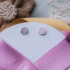 A pair of silver textured stud earrings displayed on a pink fabric surface.