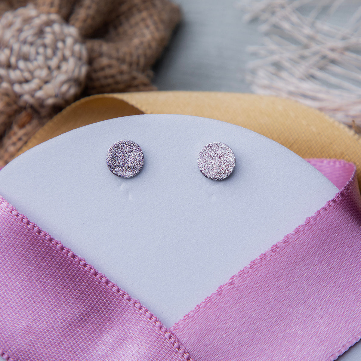 A pair of silver textured stud earrings displayed on a pink fabric surface.