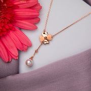 A rose gold pendant chain featuring a clover-shaped design with a suspended pearl, displayed against a floral background.