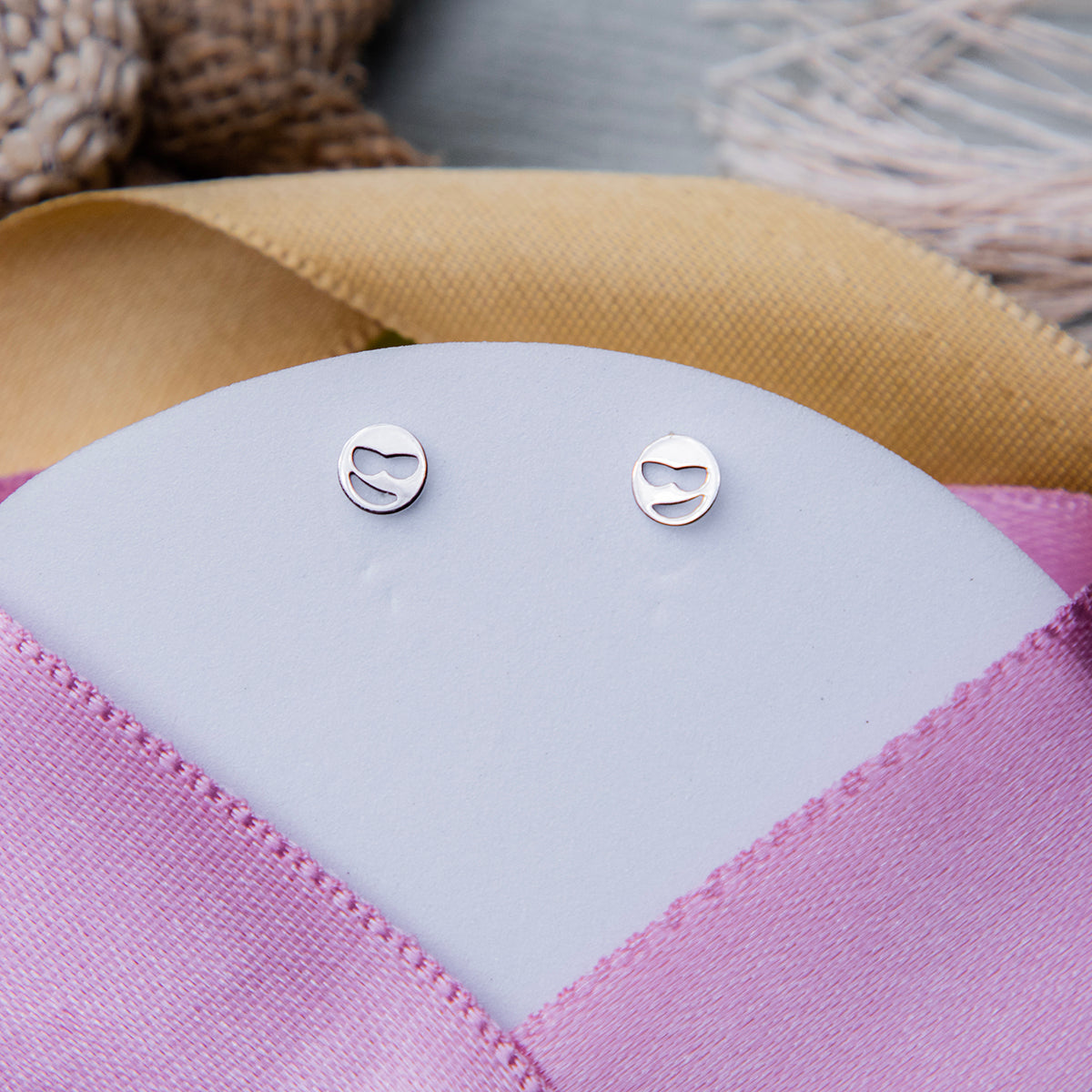A pair of silver-colored stud earrings with a cool boy emoji design, displayed on a pink cushion.