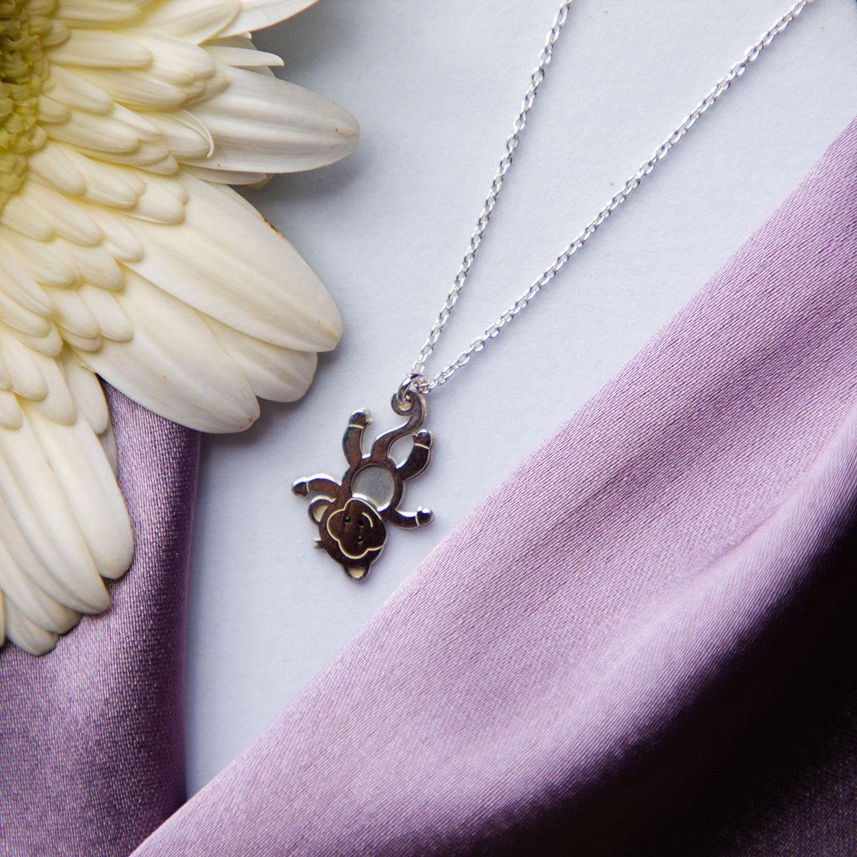 A sterling silver pendant chain featuring a monkey motif, displayed alongside a white flower on a purple fabric background.