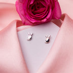 A pair of silver stud earrings designed to resemble bunny faces, displayed on a pink surface with a rose flower in the background.