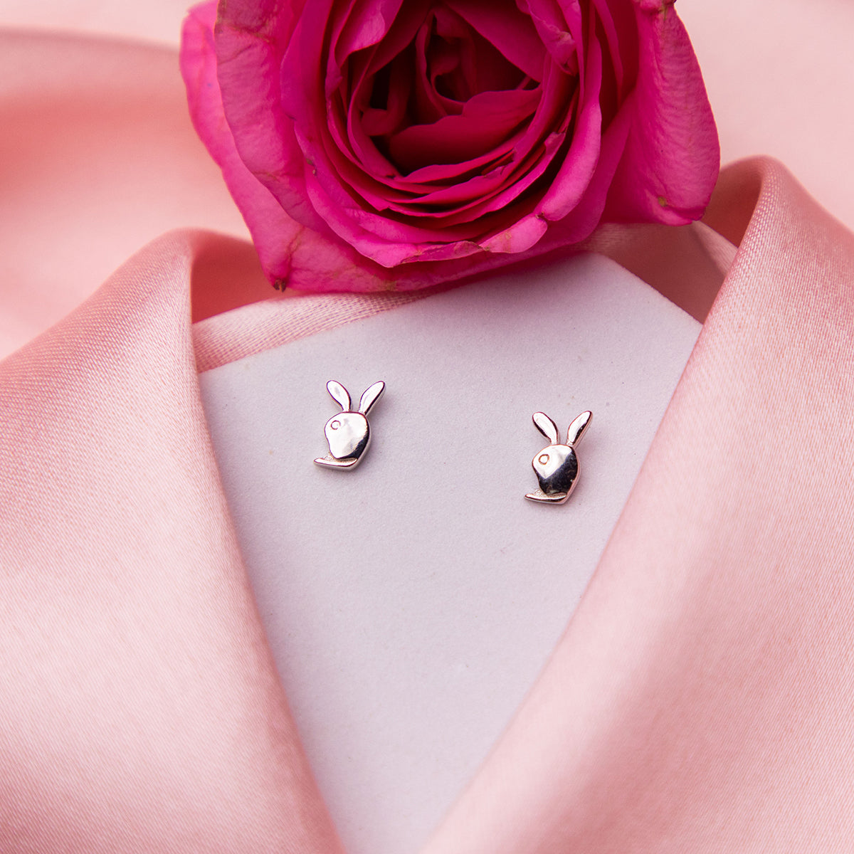 A pair of silver stud earrings designed to resemble bunny faces, displayed on a pink surface with a rose flower in the background.