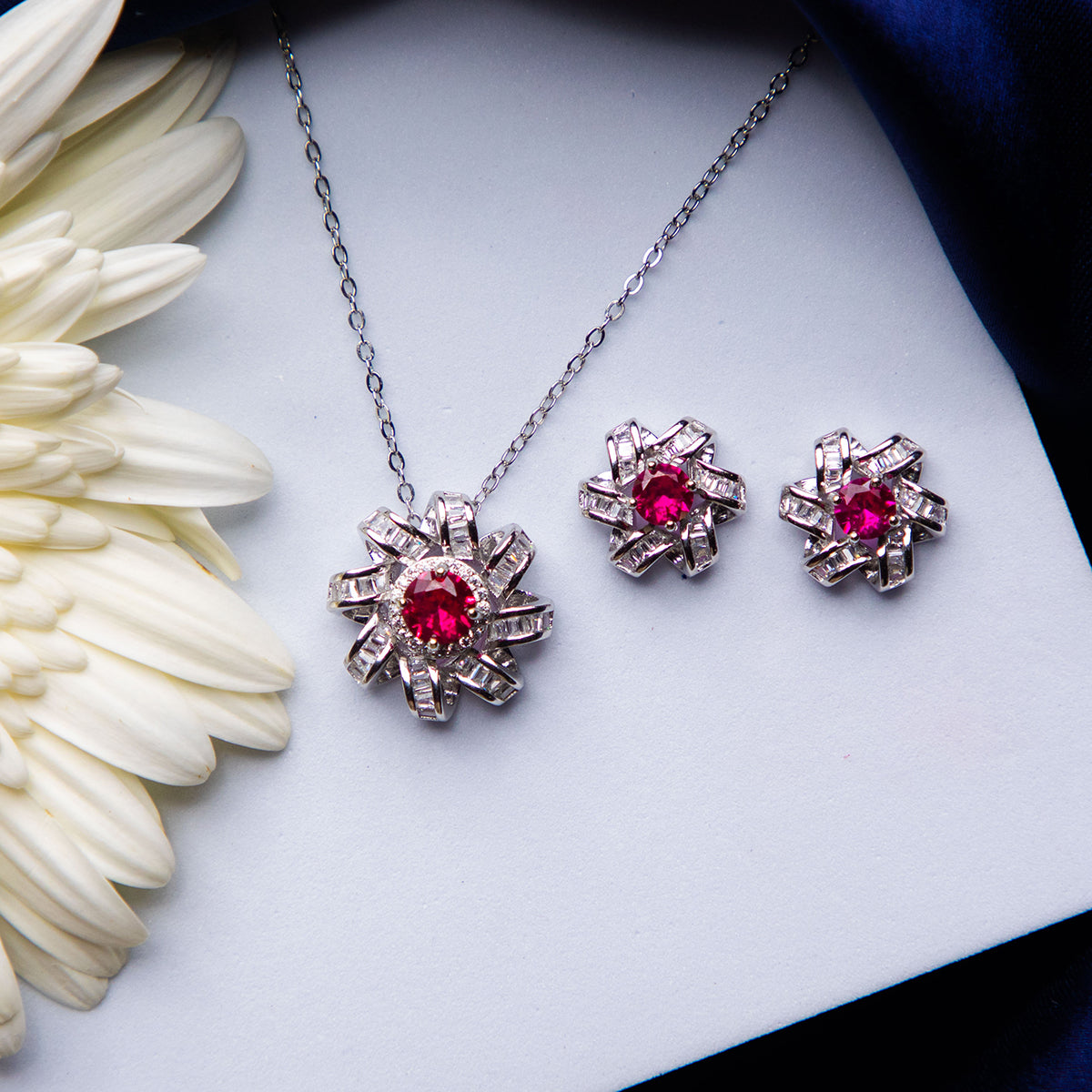 A set of jewelry featuring a dazzling pink stone pendant and a pair of earrings, placed on a white surface next to a white flower.