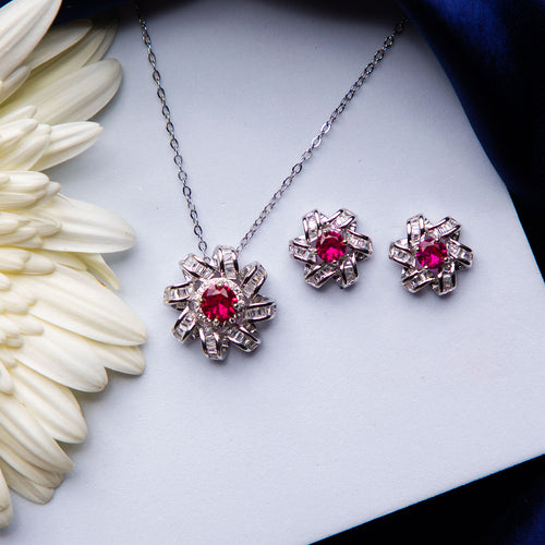 A set of jewelry featuring a dazzling pink stone pendant and a pair of earrings, placed on a white surface next to a white flower.