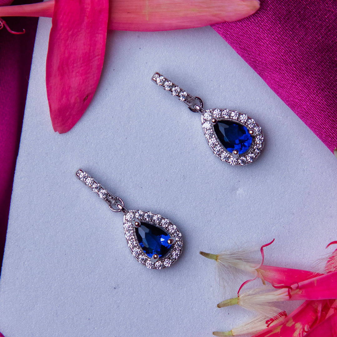 A pair of sterling silver drop earrings with deep blue gemstones and white accents, displayed on a surface with pink and blue floral accents.