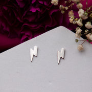 A pair of silver stud earrings designed to resemble an electric shock, displayed on a white surface with a flower and petals in the background.