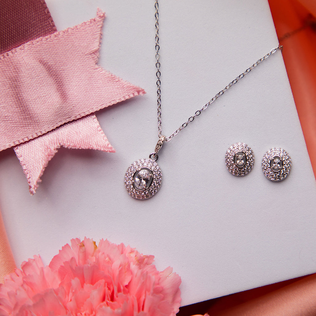 A set of sterling silver jewelry including a pendant and a pair of earrings, displayed on a white surface with a pink and red ribbon background and a flower beside it.