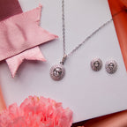 A set of sterling silver jewelry including a pendant and a pair of earrings, displayed on a white surface with a pink and red ribbon background and a flower beside it.