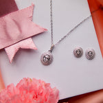 A set of sterling silver jewelry including a pendant and a pair of earrings, displayed on a white surface with a pink and red ribbon background and a flower beside it.
